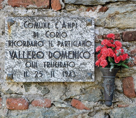 Lapide in marmo su muro di pietra, con iscrizione e vaso di fiori rossi accanto.
