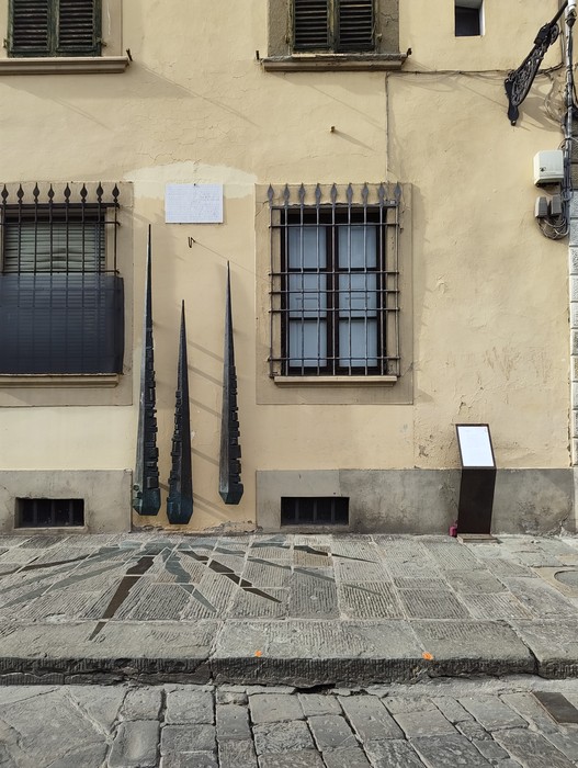 Tre sculture in ferro a forma di cipresso appese alla facciata di un edificio, accanto a una targa.
