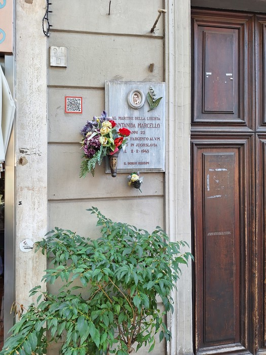 Lapide in marmo con iscrizione su una parete esterna, accanto a un portone, con un mazzo di fiori appeso sotto.