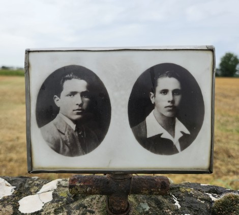 Cippo commemorativo con fotografie in bianco e nero di due giovani su sfondo di campi coltivati.