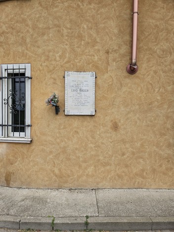 Lapide commemorativa su un muro di color ocra, con fiori e dedica a Ruggeri.