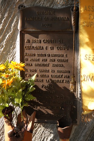 Targa in bronzo con iscrizione dedicata ai caduti di Cefalonia e Corfù, fissata alla roccia con fiori sotto.
