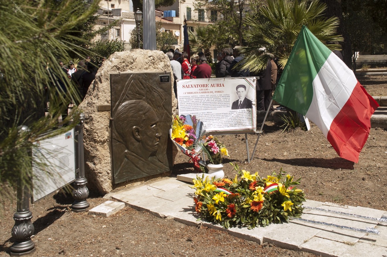 Monumento a Auria, commemorazione con bandiera italiana e fiori in un giardino pubblico.