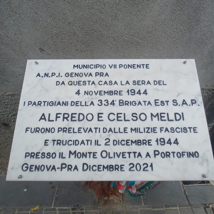 Lapide di marmo con iscrizione, fissata a una parete esterna intonacata.