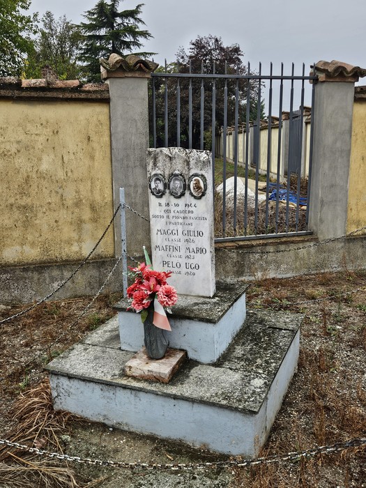 Cippo in pietra con iscrizione e foto dei caduti, posto su basamento, con fiori davanti a una cancellata.