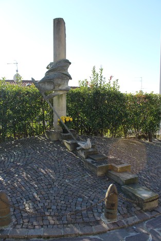 Monumento alla Resistenza con statua e corda, circondato da siepi e pavimento in pietra.