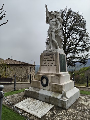 Monumento ai caduti con statua e dedica commemorativa in un paesaggio verde e collinare.