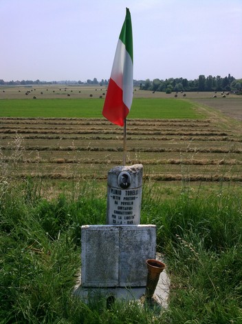 Cippo commemorativo dedicato a Plinio Torelli, con bandiera italiana, immerso in un paesaggio agricolo.
