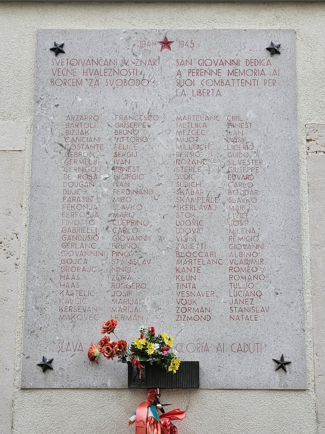 Lapide commemorativa in onore dei caduti per la libertà, con nomi incisi e decorazioni floreali.