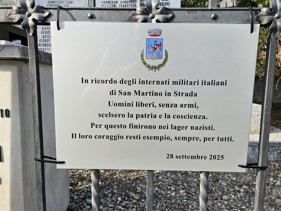 Targa bianca con stemma comunale e iscrizione, fissata a un cancello in ferro battuto nei pressi di un cimitero.