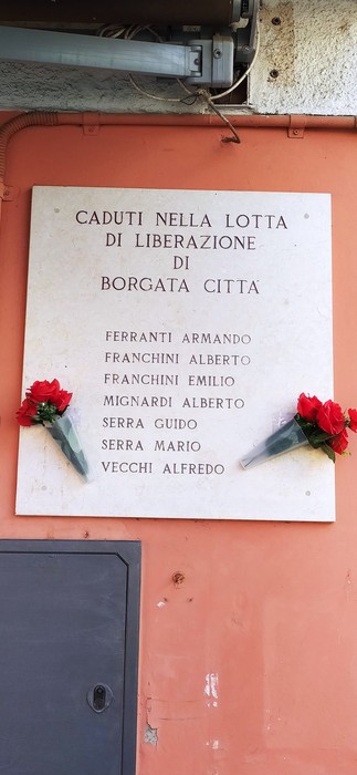 Lapide commemorativa dedicata ai caduti della lotta di liberazione nella Borgata Città. Fiori rossi adornano la pietra.