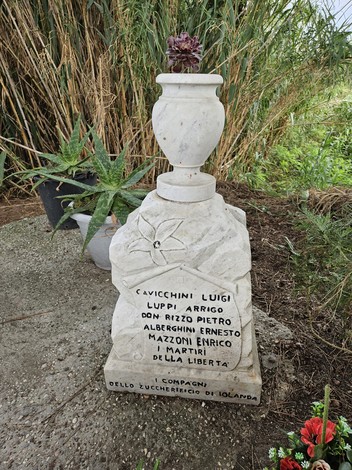 Monumento ai Martiri della Macchinina con iscrizioni e fiori, immerso in un contesto naturale.