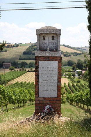 Cippo commemorativo dedicato a Baldini e Raimondi, circondato da un paesaggio di colline e vigneti.