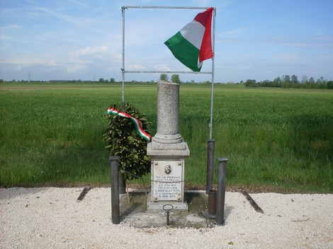 Cippo commemorativo a Battini, con bandiera italiana e decorazioni florali, immerso in un paesaggio verde.